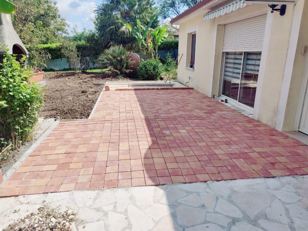 Terrasse en pavés rouges et beiges devant une maison crème, avec un jardin en terre labourée et végétation luxuriante en fond.