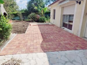 Terrasse en pavés rouges et beiges devant une maison crème, avec un jardin en terre labourée et végétation luxuriante en fond.