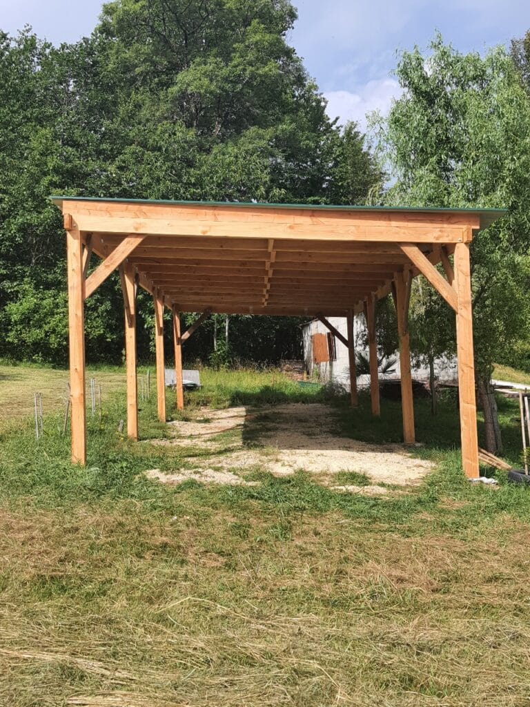 Grand carport en bois clair à toit vert sur une allée de graviers, entouré de verdure et d'arbres sous un ciel bleu.