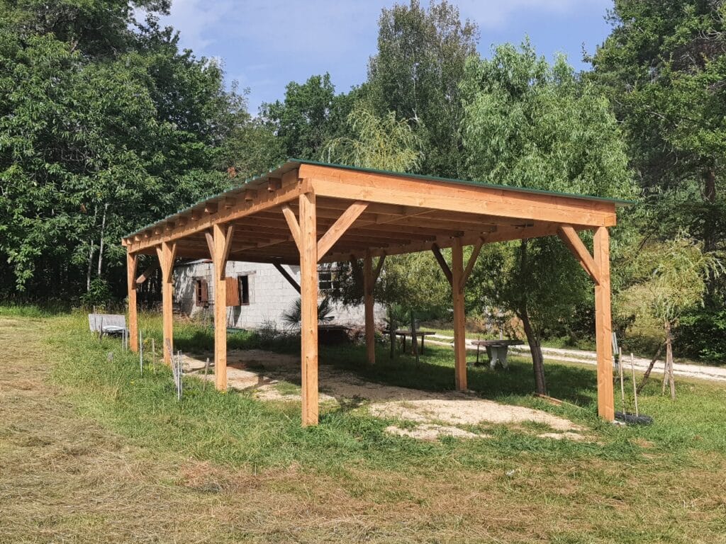 Grand carport en bois clair à toit vert ondulé, dans un cadre naturel arboré. Un bâtiment en parpaings est visible derrière.