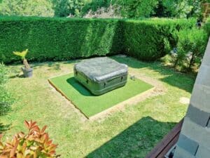 Jacuzzi extérieur noir couvert sur gazon artificiel, dans un jardin verdoyant avec haie taillée et palmier en pot.