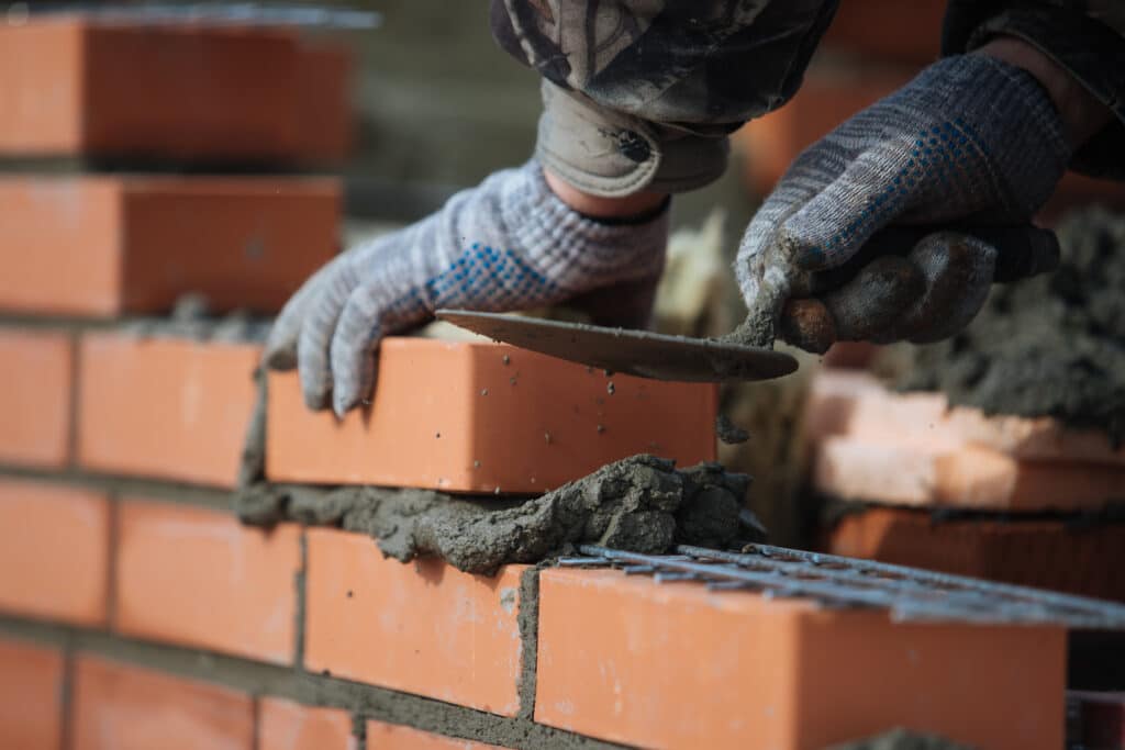 Mains gantées d'un maçon posant des briques orange avec du mortier et de l'armature métallique pour construire un mur.