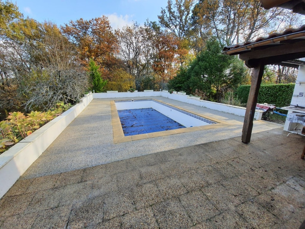 Piscine extérieure couverte d'une bâche bleue, entourée d'une terrasse pavée et d'arbres aux couleurs automnales. Ambiance de jardin paisible.