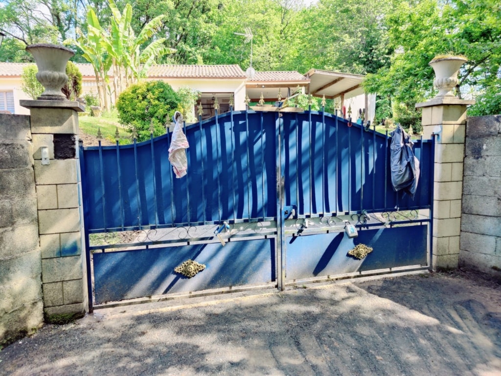 Grand portail bleu en métal double battant, flanqué de piliers en pierre avec urnes décoratives. L'entrée s'ouvre sur une propriété verdoyante avec arbres et maison en arrière-plan. Un sac et un tissu sont accrochés au portail.