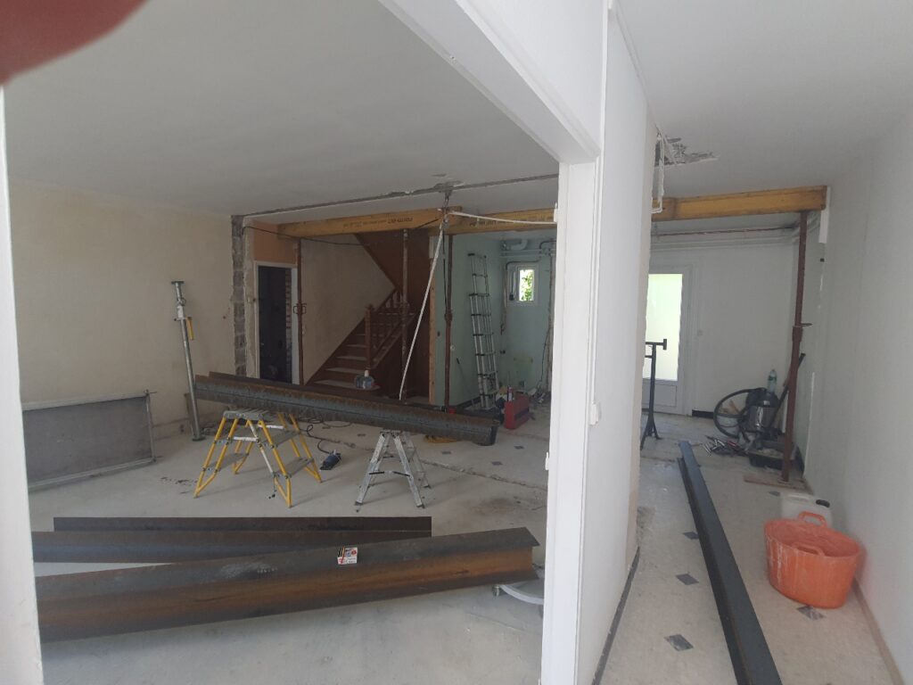 Intérieur de maison en rénovation structurelle. Des poutres métalliques et étais temporaires supportent le plafond. Murs démolis, escalier visible.
