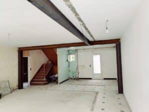 Salle de vie en rénovation avec poutres IPN et poteaux métalliques apparents, escalier bois, murs dénudés et sol en béton.