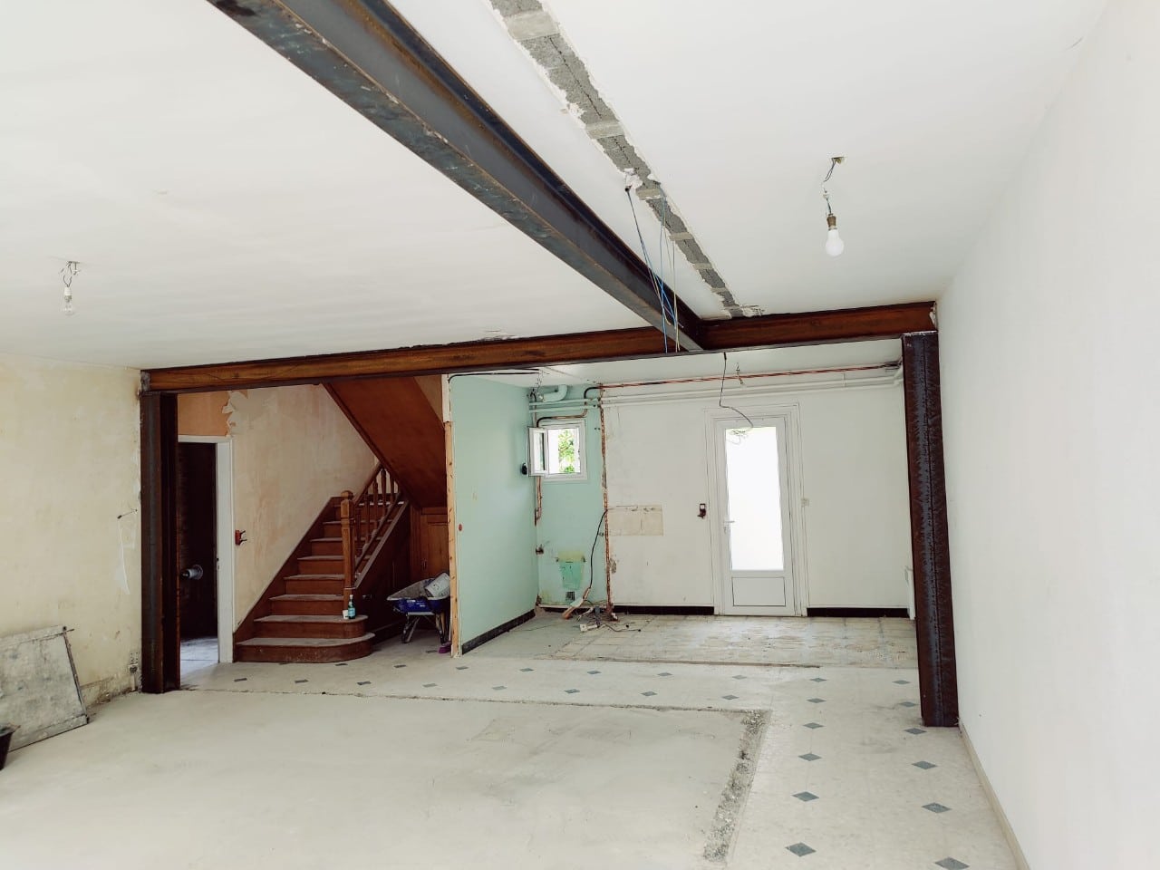 Salle de vie en rénovation avec poutres IPN et poteaux métalliques apparents, escalier bois, murs dénudés et sol en béton.
