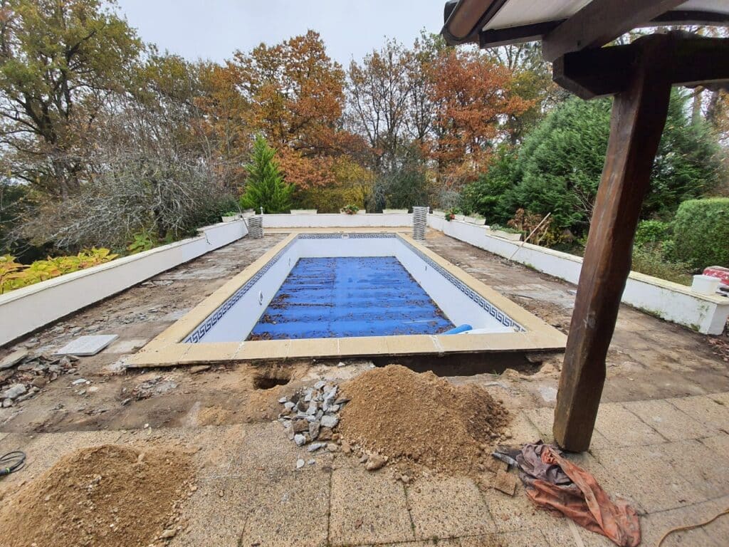 Piscine vide en rénovation avec bâche bleue, terrasse en travaux, et arbres aux couleurs d'automne en arrière-plan.