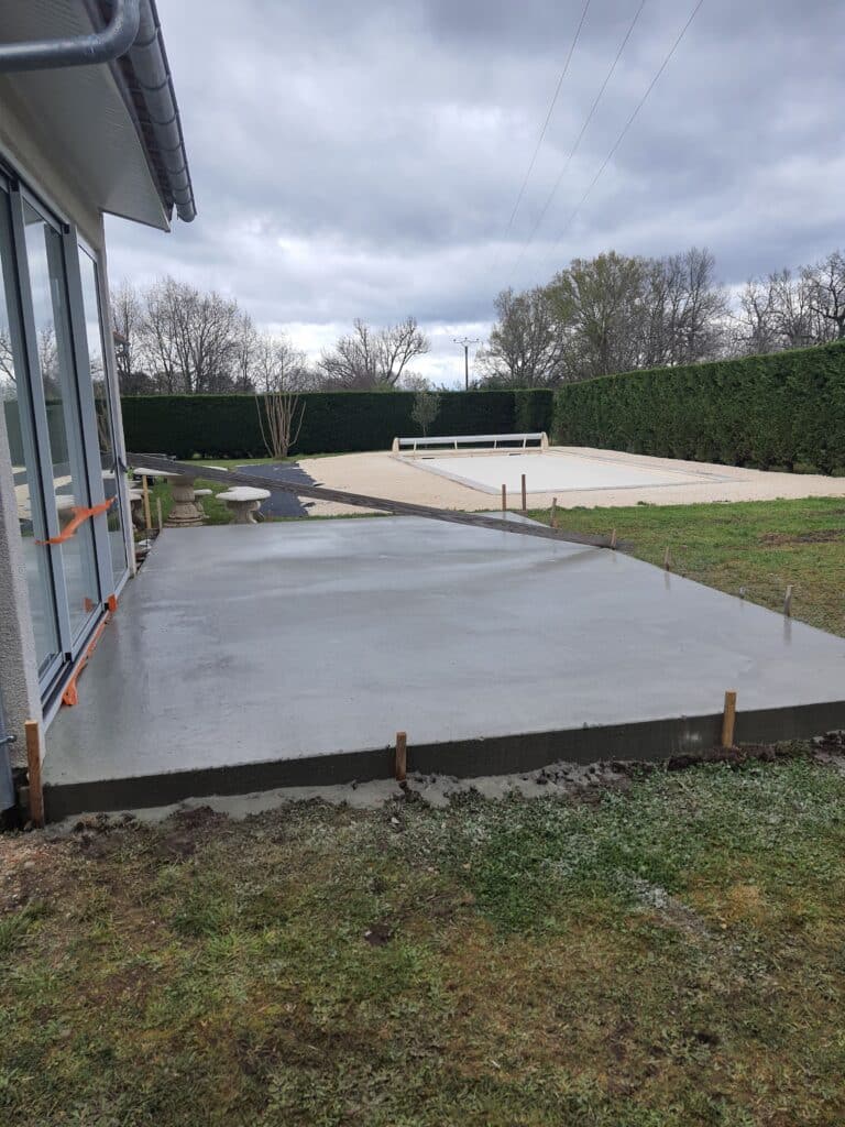 Terrasse en béton fraîchement coulée devant une maison, avec jardin, haie verte et zone de gravier en arrière-plan sous un ciel nuageux.