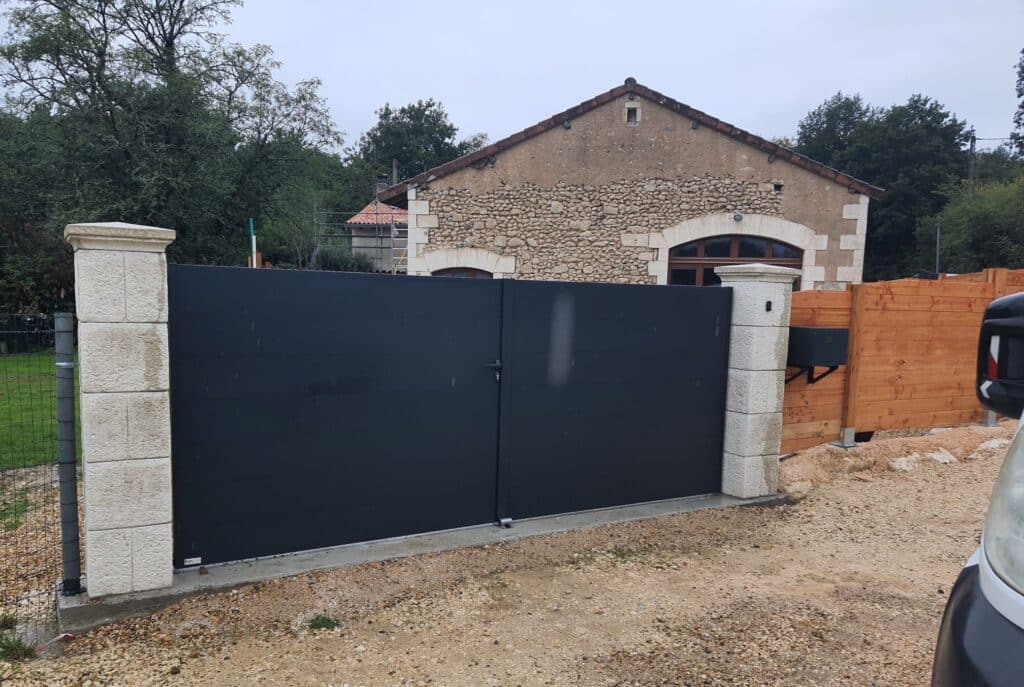 Portail battant en alu gris foncé entre deux piliers de pierre. À l'arrière, une maison ancienne en pierres rustiques. Boîte aux lettres 13.