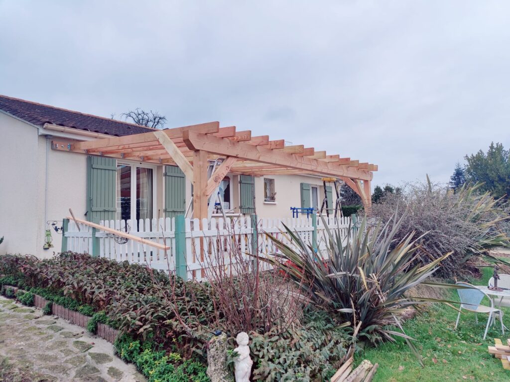 Pergola bois en construction adossée à une maison aux volets verts avec terrasse et clôture blanche de jardin.